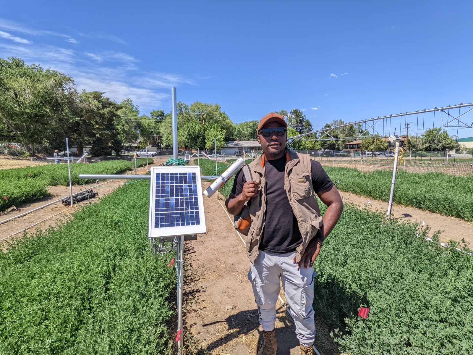 Emmanuel Fru standing in a green test field beside a small solar panel under a blue sky